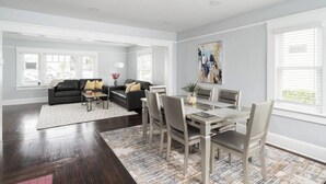 A spacious living room with a dark wood floor, a black leather sofa with beige cushions, a glass dining table with six chairs, and a large abstract painting on the wall.