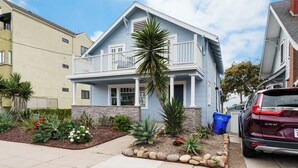A two-story house with a balcony, a stone foundation, and a well-maintained garden with various plants and a palm tree.