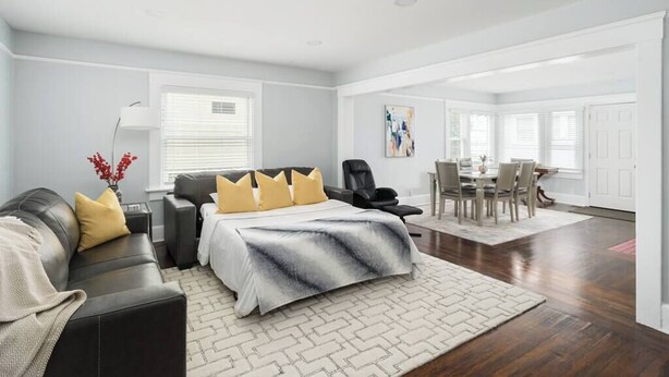 A modern living room with a sofa, a dining table, and chairs. There is a bed with a white and gray bedspread.