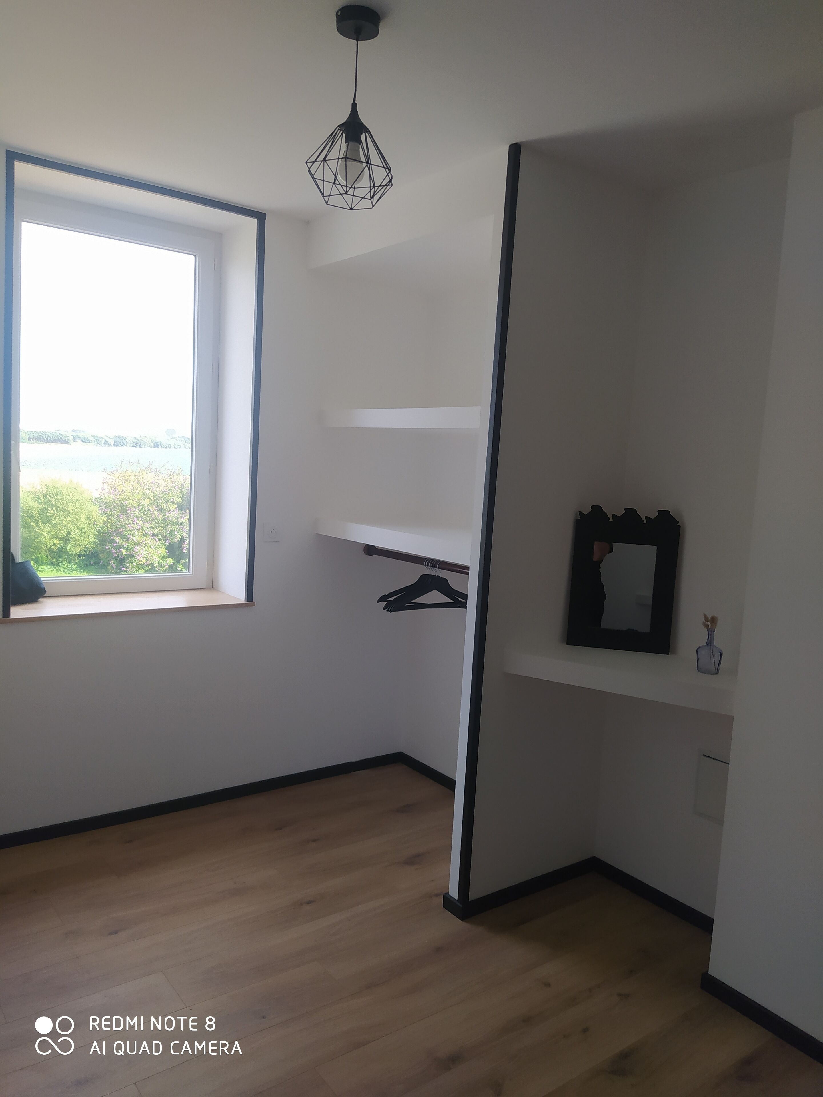 Een kamer met raam, houten vloer, hanglamp, spiegel en een kleine vaas.