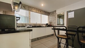 Een moderne keuken met witte kastjes, een zwart aanrecht en een raam. Er zijn twee barkrukken en een koelkast.