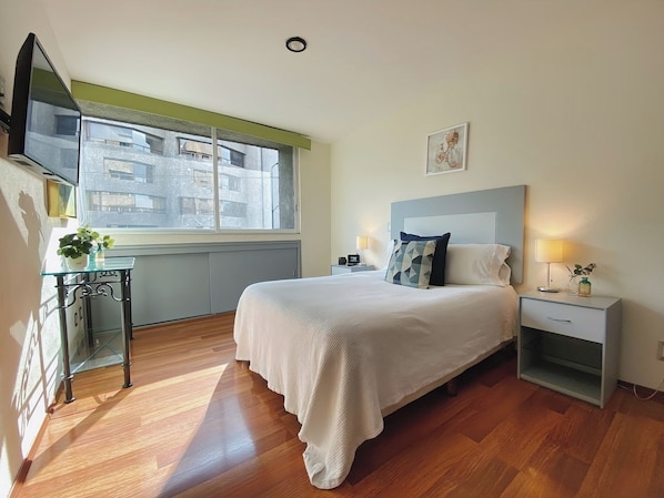 A hotel room with a bed, bedside tables, a TV, a window, and a view of buildings outside.