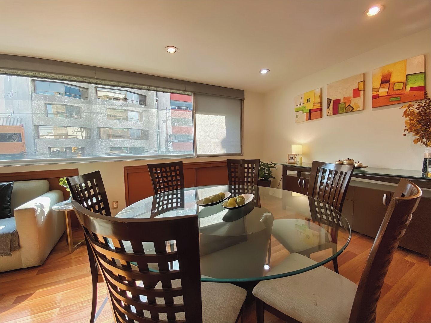 A modern dining area with a glass table and wooden chairs, a sofa, and a view of buildings through a large window.