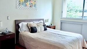 En pænt redt seng med et mønstret sengetæppe, et sengebord med en lampe og en dekorativ vægdekoration.