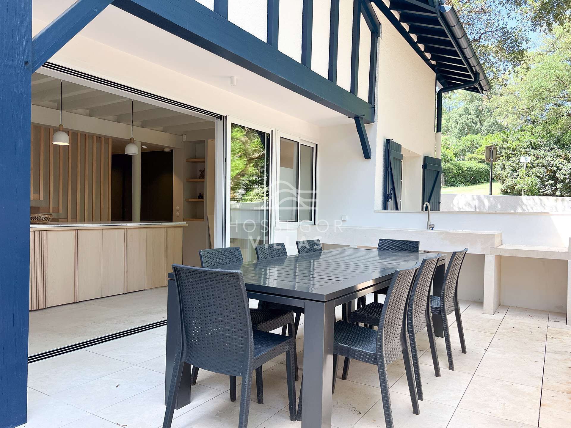 A modern outdoor dining area with a glass table and wicker chairs, connected to a kitchen with wooden cabinets and a built-in oven.
