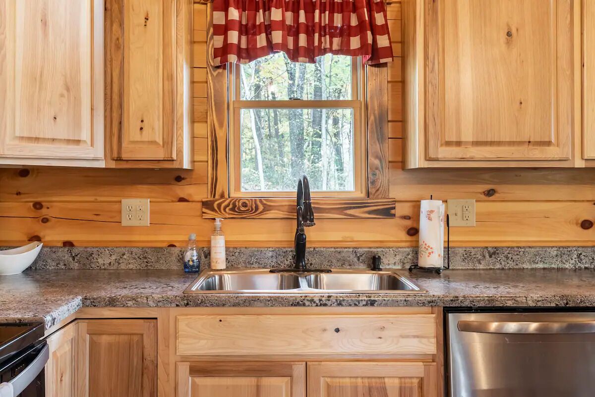 A kitchen with wooden cabinets, a granite countertop, a stainless steel sink, and a window with a view of trees.