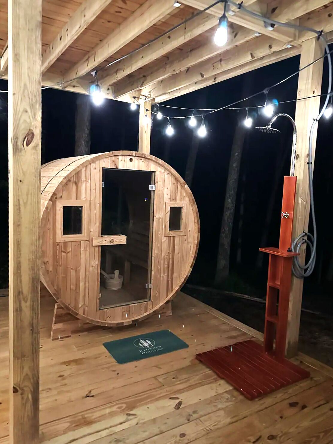 Une structure de sauna en bois, dotée d’une porte et d’un espace douche, le tout agrémenté de guirlandes lumineuses.