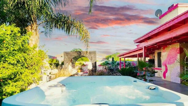 A hot tub surrounded by lush greenery and a colorful building with a pink facade. - Welcome to The Pink Cabana, where modern Moroccan meets luxe desert vibes. (Indio)