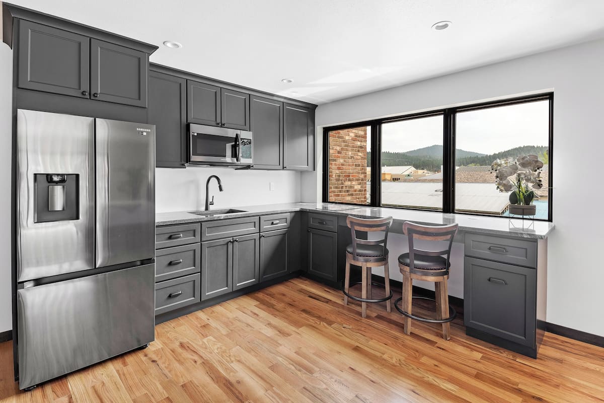 Une cuisine moderne avec des meubles gris, des appareils en acier inoxydable, un plancher en bois et une grande fenêtre donnant sur un bâtiment en briques et des collines.
