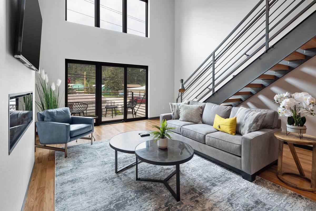 Un salon moderne avec un canapé gris, un fauteuil bleu et une table basse agrémentée d’une plante. On aperçoit un escalier au fond de la pièce.