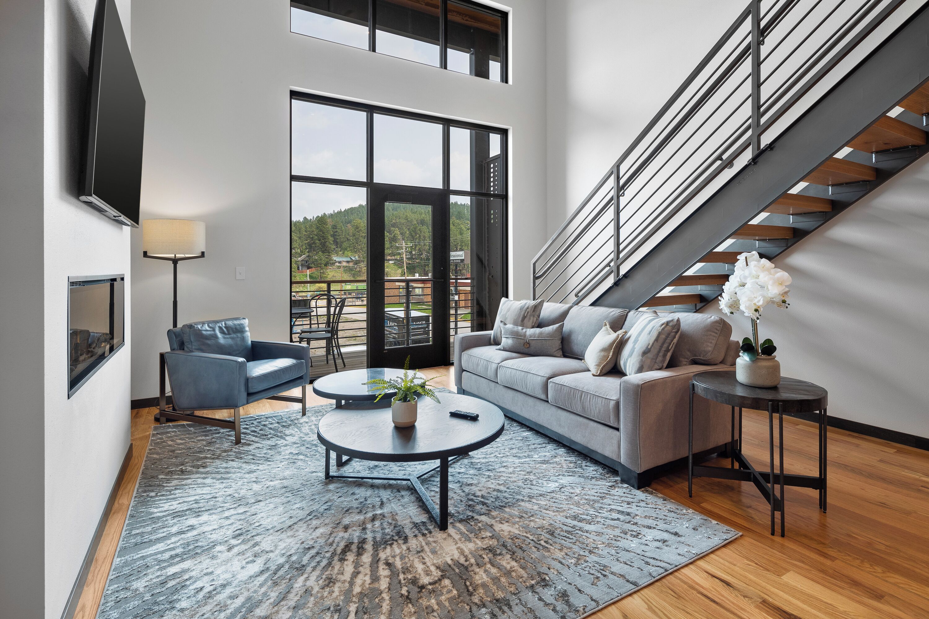 A modern living room with a grey sofa, a coffee table, a TV mounted on the wall, and a staircase leading to an outdoor area.