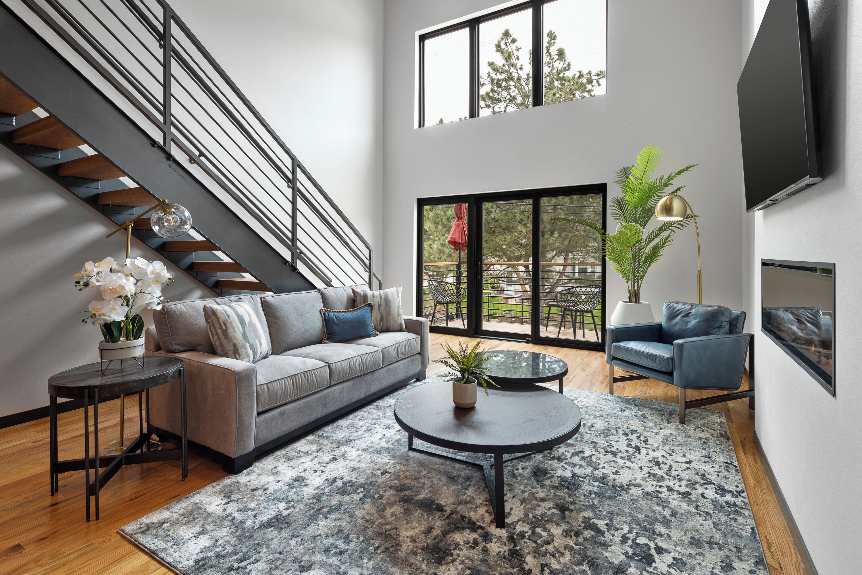 A modern living room with a grey sofa, a round coffee table, a staircase, and a large window with a view of the outdoors.