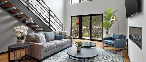 A modern living room with a grey sofa, a round coffee table, a staircase, and a large window with a view of the outdoors.