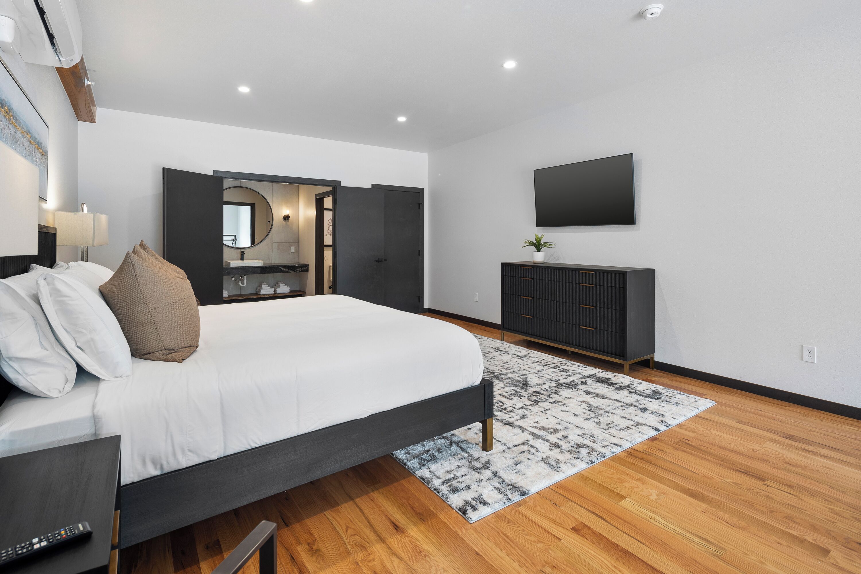 A modern bedroom with a large bed, a vanity with a mirror, a flat-screen TV, and a dark-colored dresser.