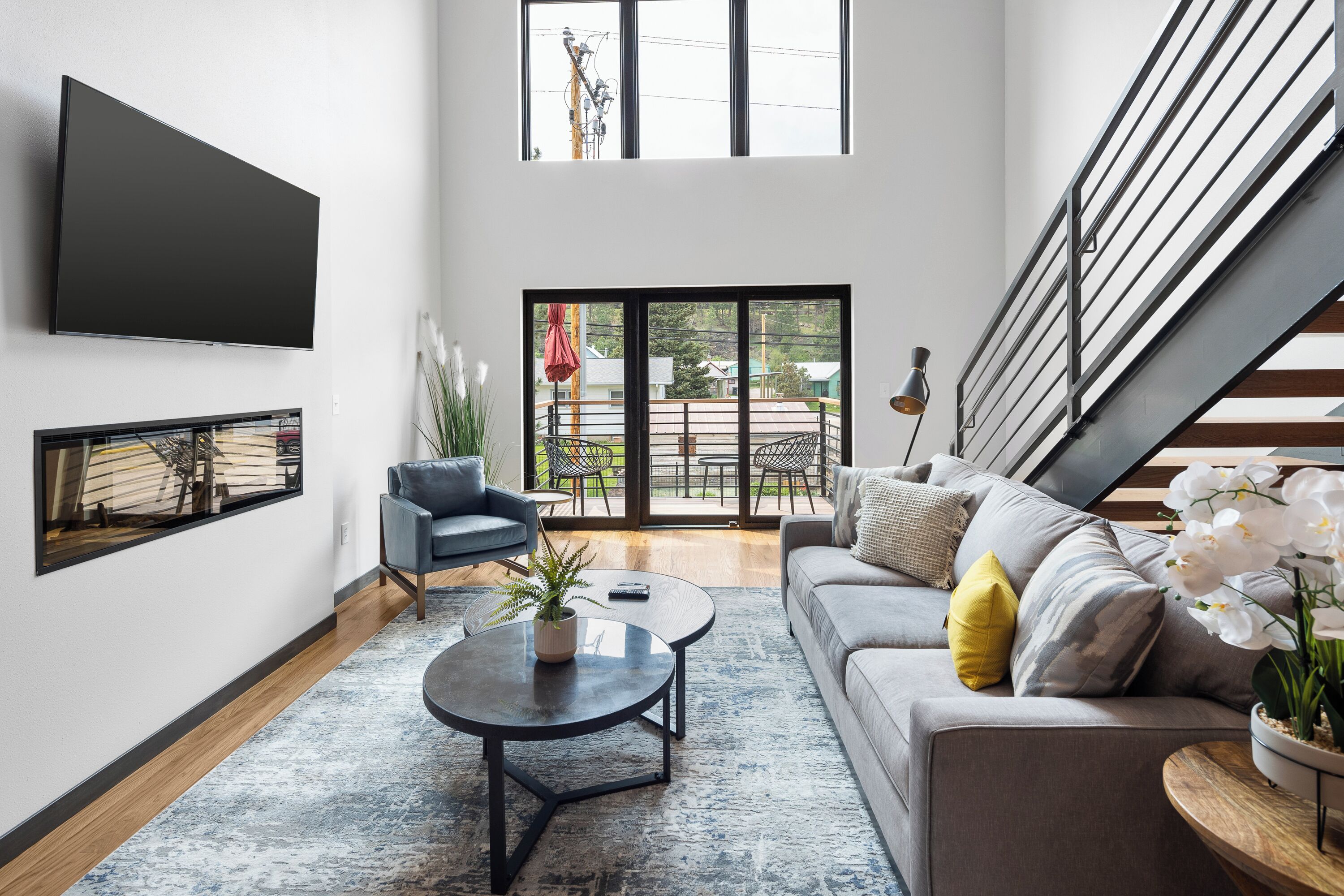 A modern living room with a large flat-screen TV, a sofa, a coffee table, and a staircase leading to an outdoor area.