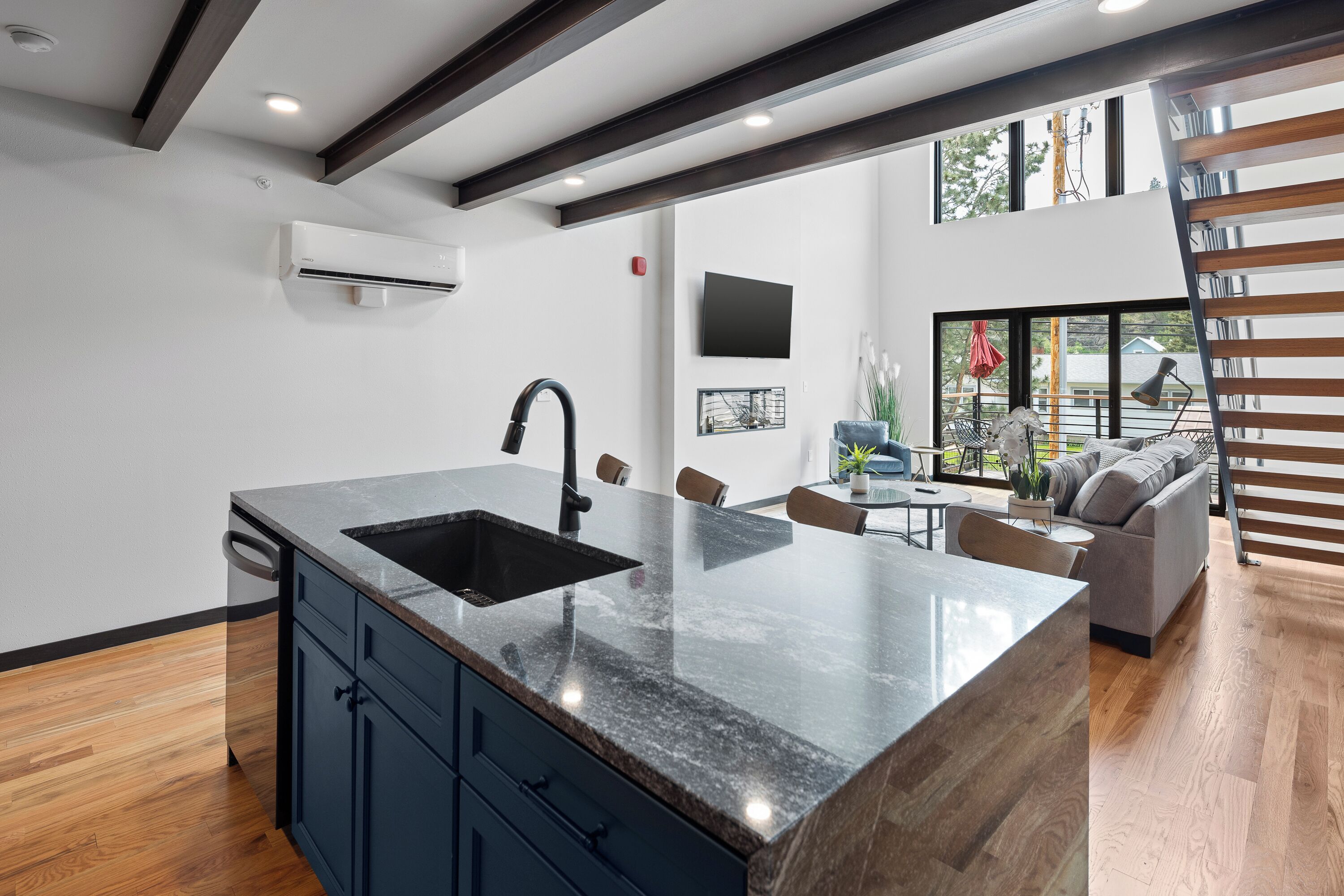 A modern kitchen with a large island, dark cabinetry, and a view to a living area with a sofa and a dining table.