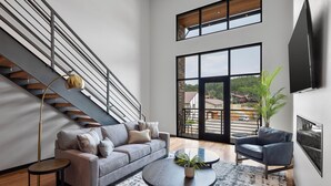 A modern living room with a grey sofa, a round coffee table, a blue armchair, a floor lamp, and a large window offering a view of the outdoors. - McGregor and Main Luxury Lofts (Hill City)