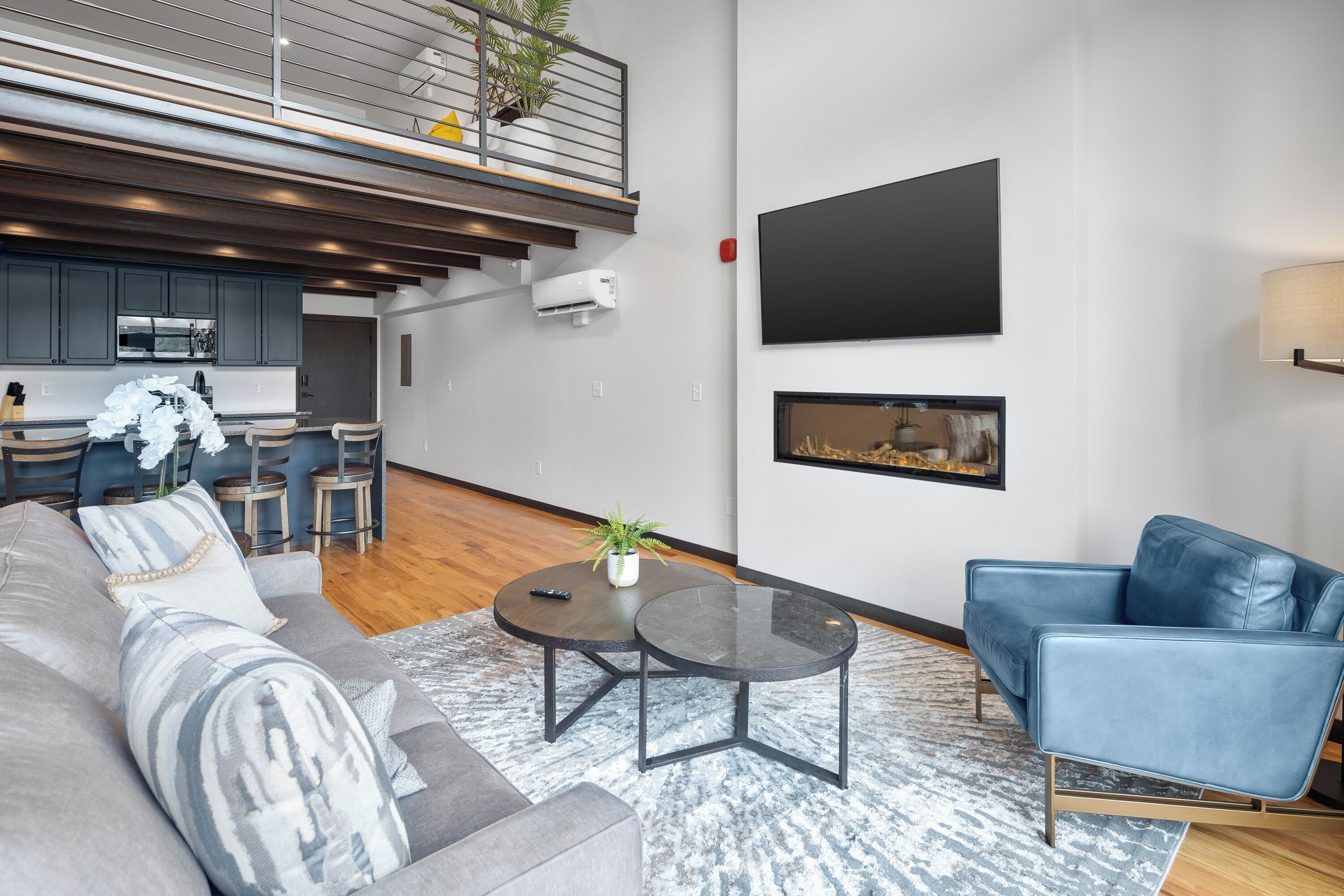 A modern living room with a grey sofa, a blue armchair, and a coffee table. There is a kitchen area in the background with grey cabinets and a wooden countertop.