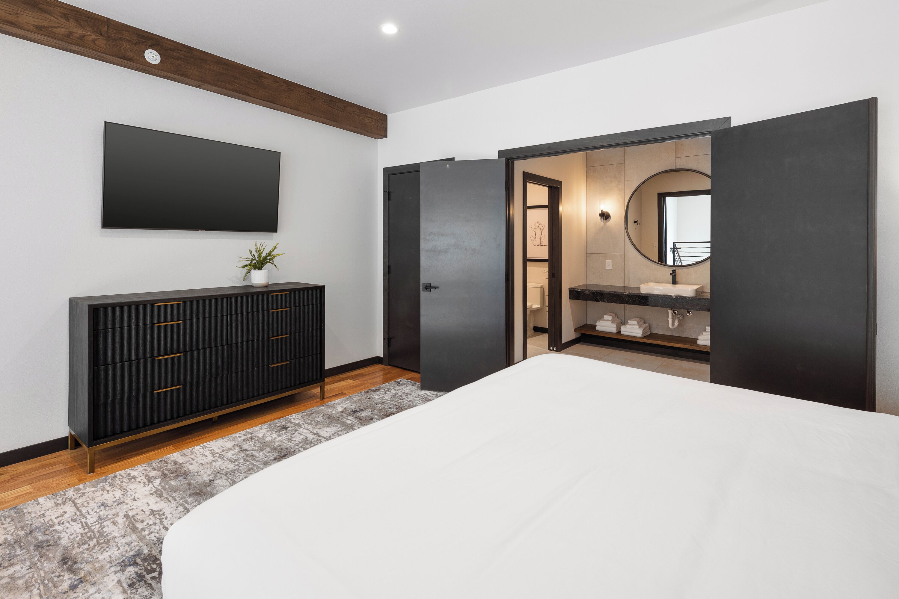 A modern bedroom with a large bed, a dark wooden dresser, a flat-screen TV, and a bathroom with a large mirror and sink.