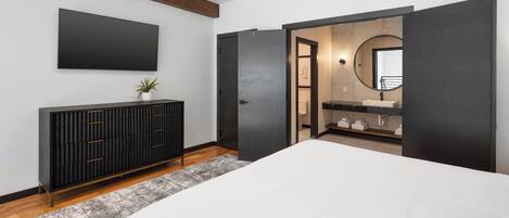 A modern bedroom with a large bed, a dark wooden dresser, a flat-screen TV, and a bathroom with a large mirror and sink.