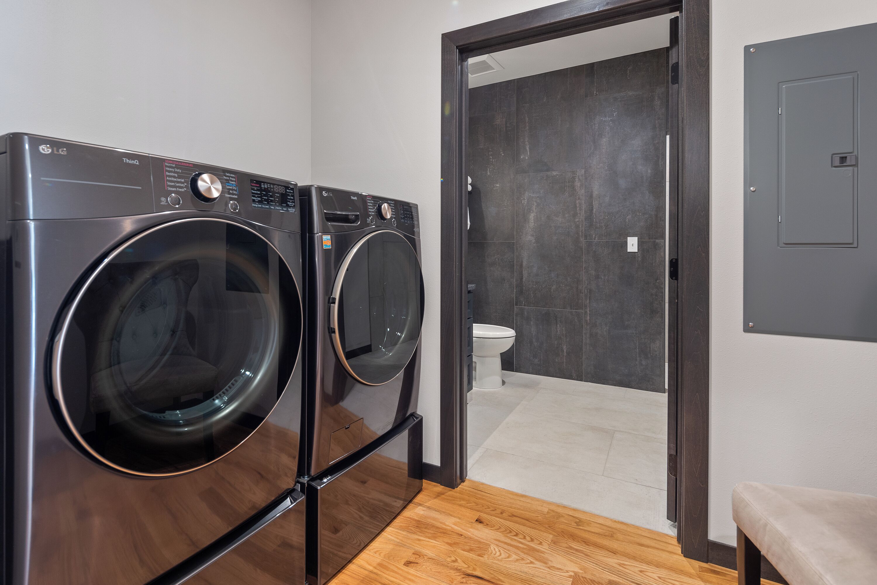 Een wasruimte met twee voorladers wasmachines en drogers, een houten vloer en uitzicht op een badkamer met toilet en donkere tegels.