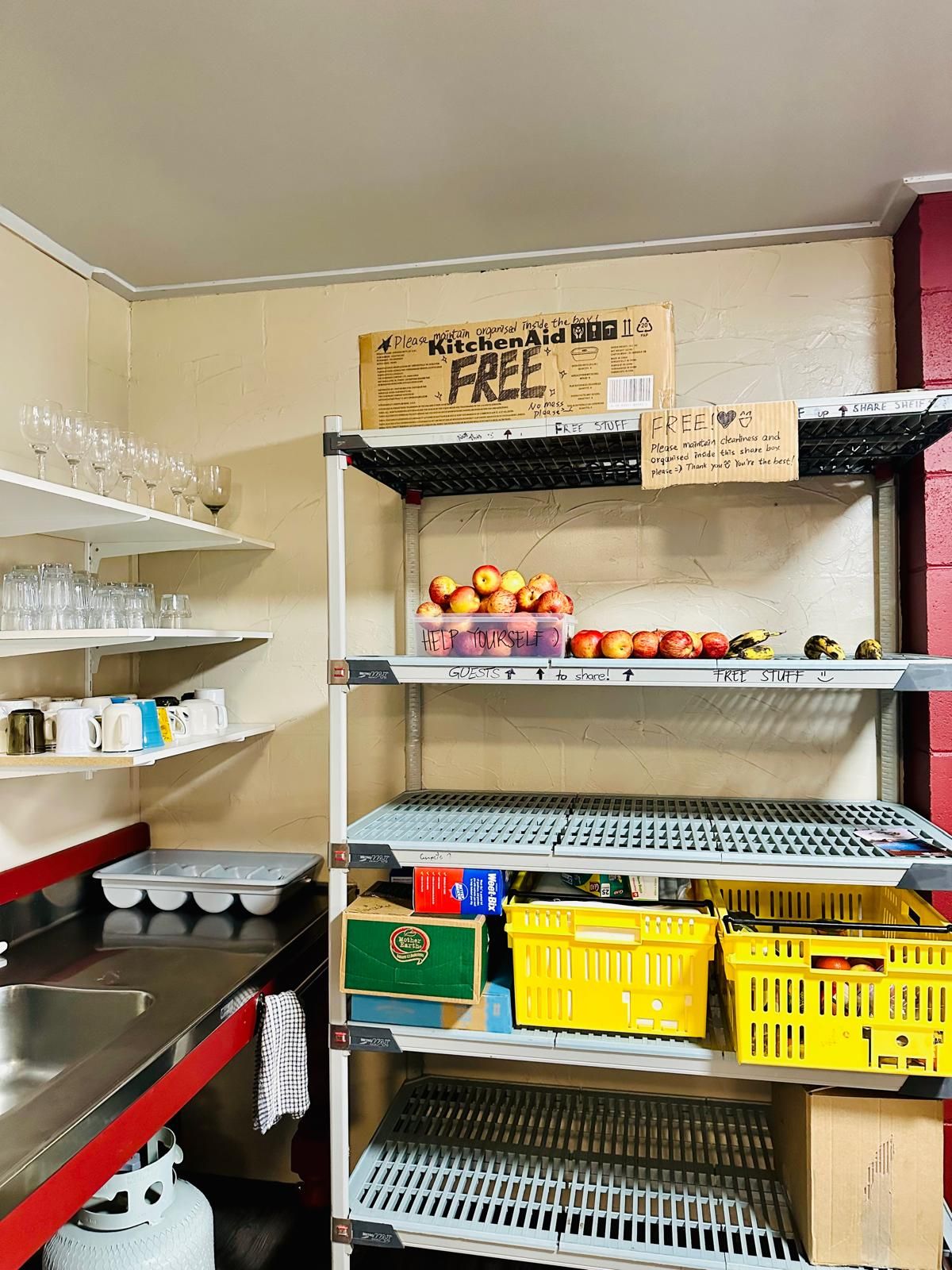 A kitchen with shelves holding various items including a box labeled 'KitchenAid FREE,' a container with apples, and several yellow crates.