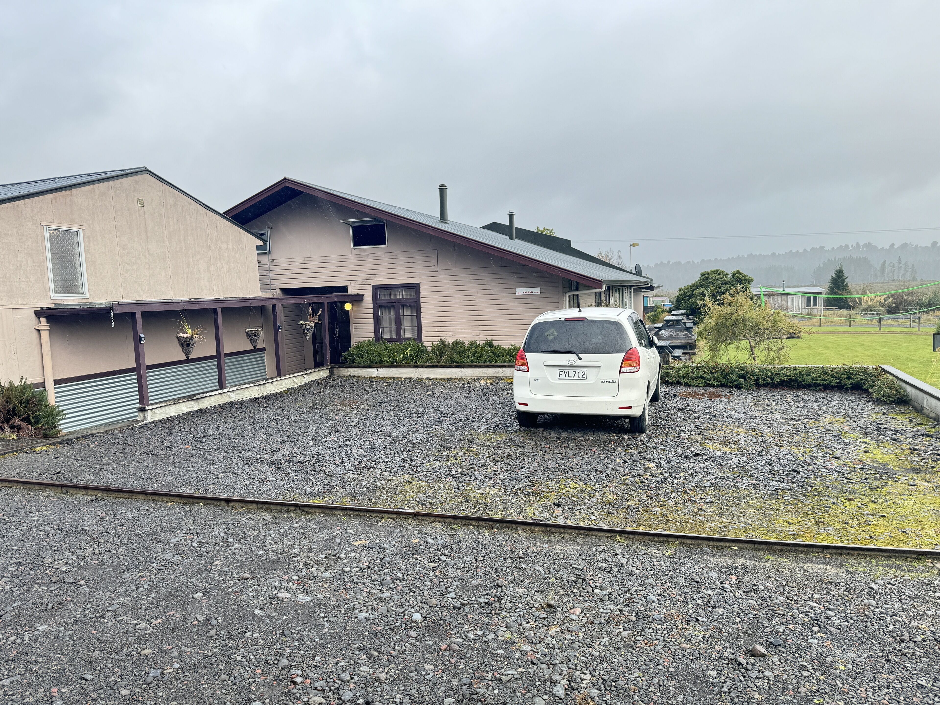 A white car parked in a gravel driveway in front of a building with a metal roof and a sign that reads "Parking."