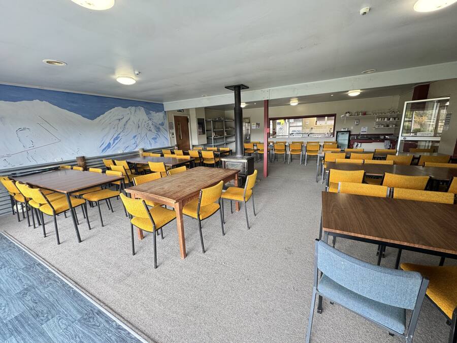 Uma área de refeições com cadeiras amarelas e mesas de madeira, um mural de montanhas na parede e uma cozinha ao fundo.