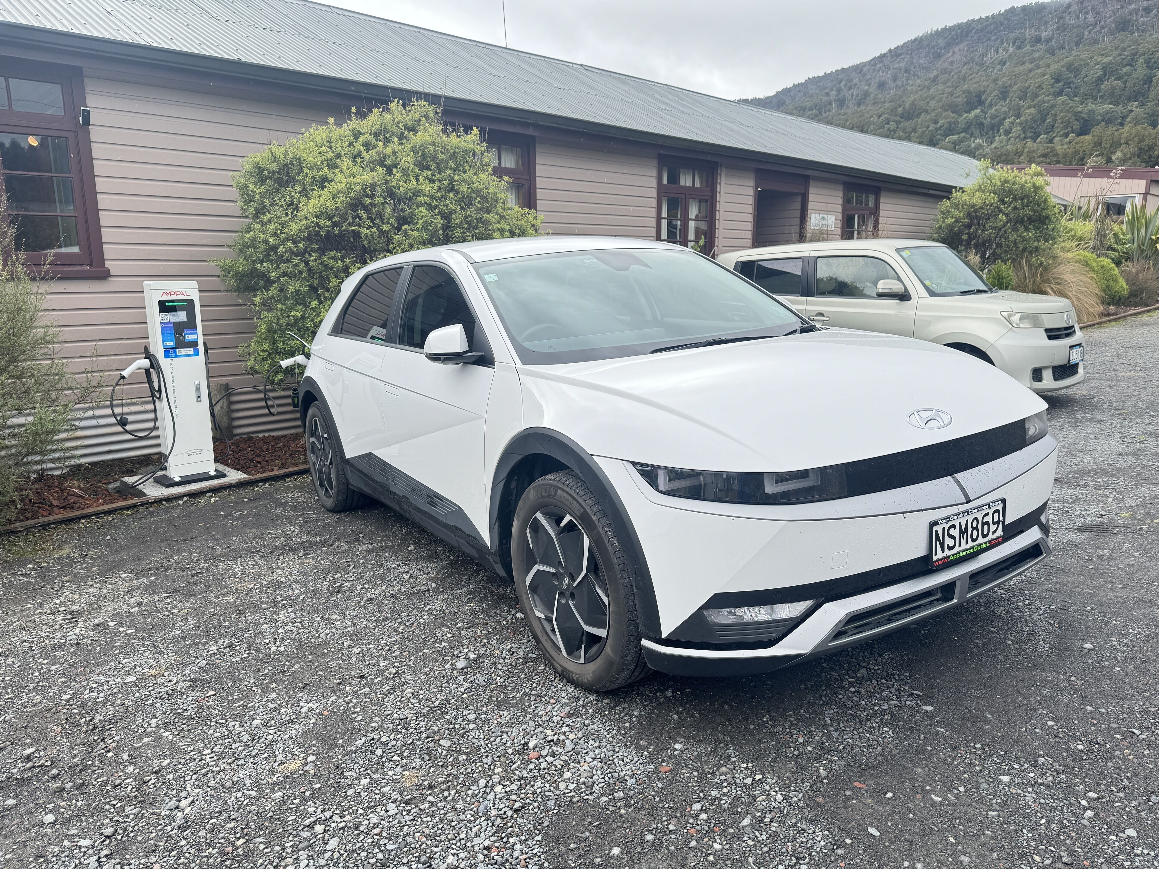 Ein weißer SUV parkt an einer Ladestation vor einem Gebäude mit einem geneigten Dach und einer Schotterauffahrt.