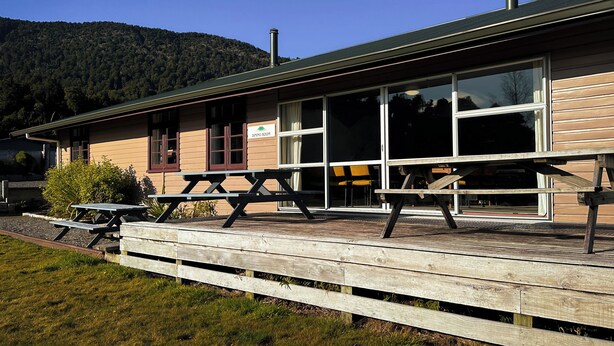 Un comedor con terraza de madera y mesas de picnic, una chimenea y montañas al fondo.