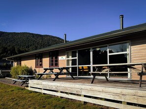 A dining room with a wooden deck and picnic tables, a chimney, and a mountain backdrop. - The Crossing Lodge (Erua)