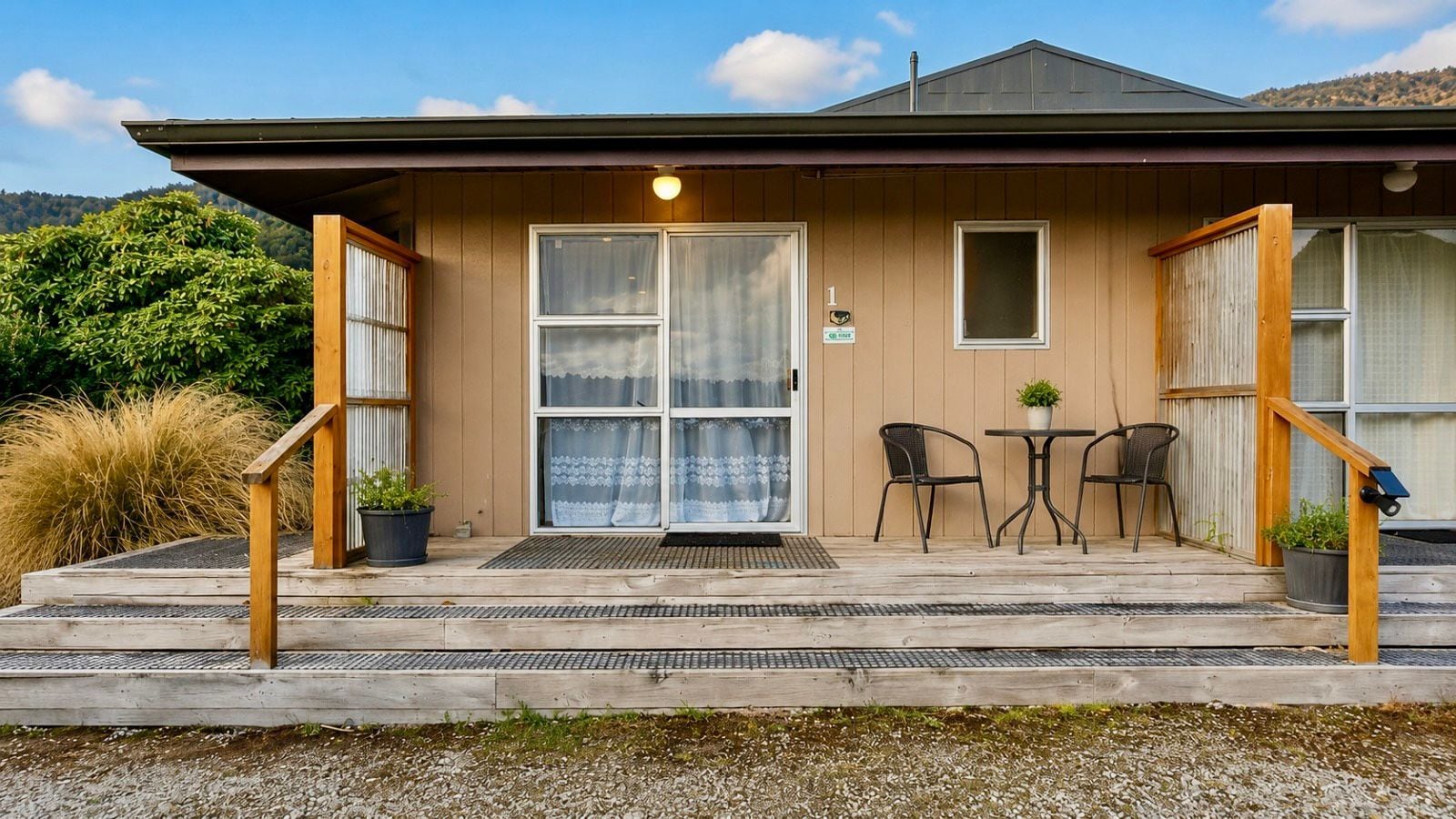 A building with a wooden exterior, a covered entrance with a door, two chairs, a table, and a small potted plant.