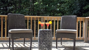 Deux chaises en osier avec des coussins, posées sur une terrasse en bois, une table décorative en métal avec un plateau à motifs et, au fond, une rambarde en bois.