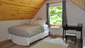 Une chambre avec un plafond en pente, un lit, un bureau et une fenêtre donnant sur un paysage verdoyant.