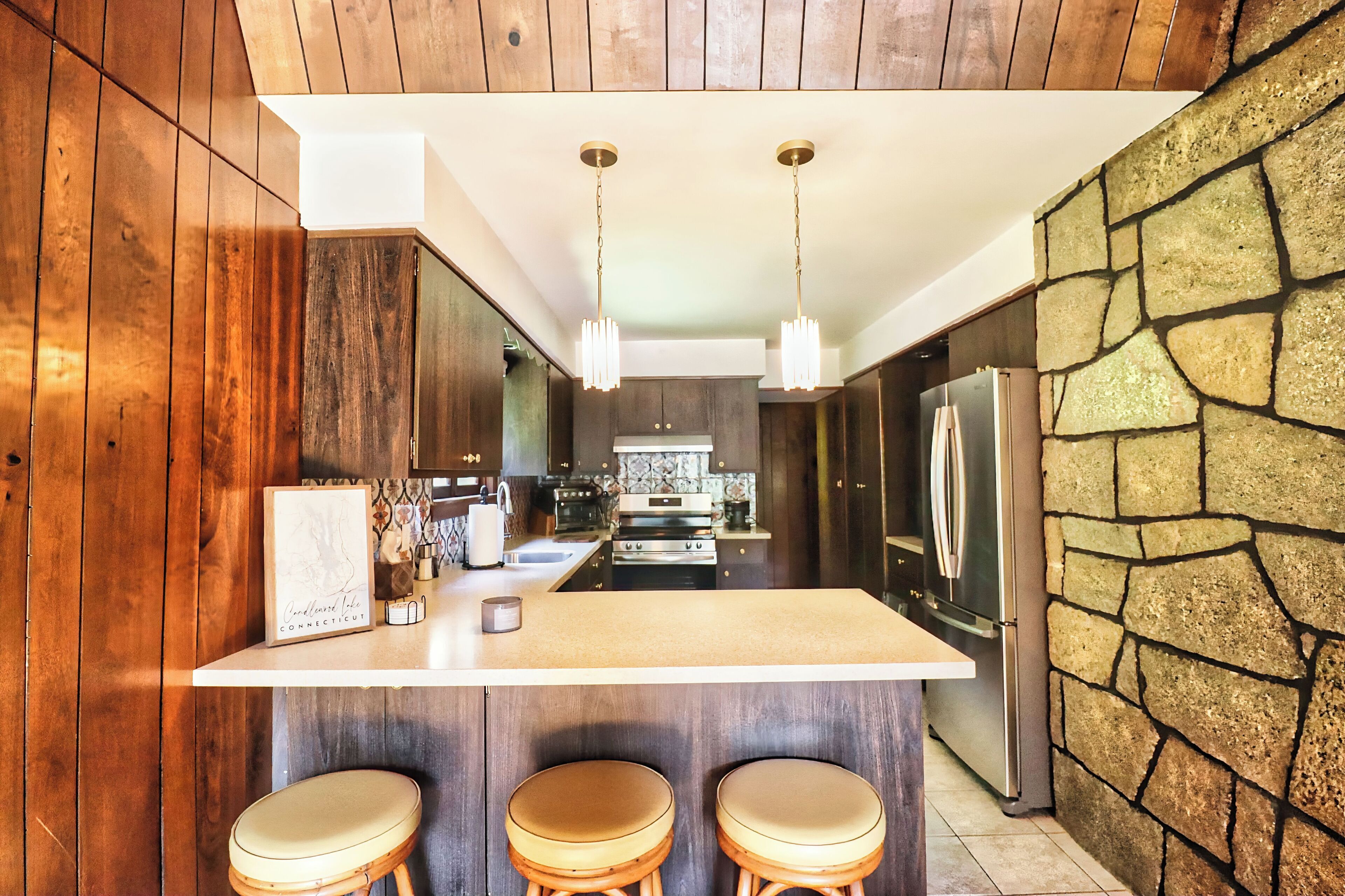 Une cuisine moderne avec un lambris en bois, un îlot central doté de tabourets, un mur d’accent en pierre et des appareils électroménagers en acier inoxydable.
