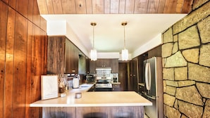 Una cocina moderna con paneles de madera, una isla central con taburetes, una pared decorativa de piedra y electrodomésticos de acero inoxidable.