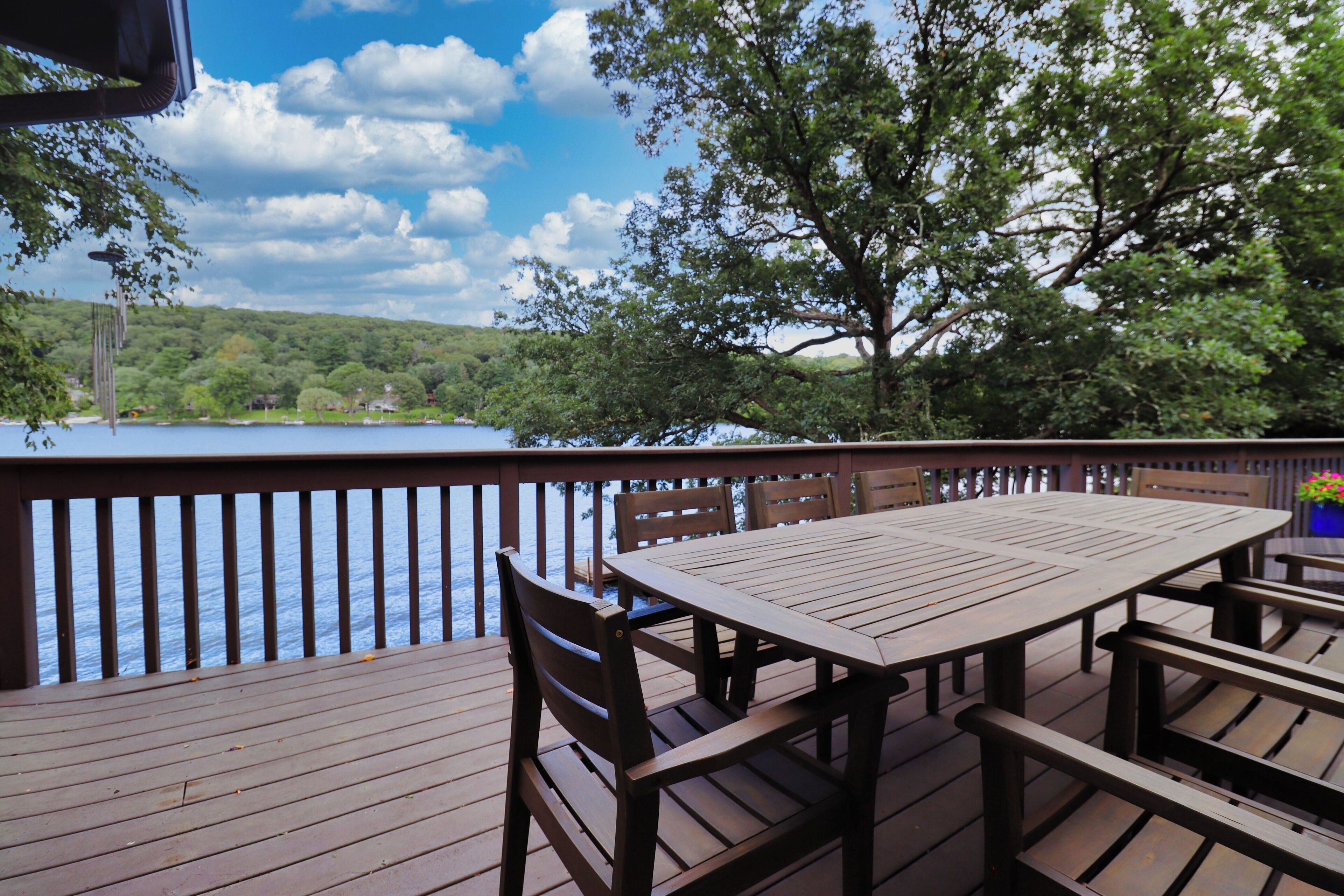 Una terraza de madera con una mesa y sillas, con vista a un lago y árboles al fondo.