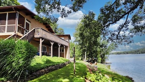 Una cabaña junto al lago con terraza de madera, porche cubierto y vistas al lago.