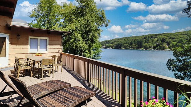 Terraza de madera con mobiliario exterior, con vista a un lago y rodeada de árboles.