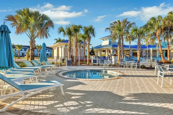A pool area with lounge chairs and umbrellas, surrounded by palm trees and buildings with blue roofs.