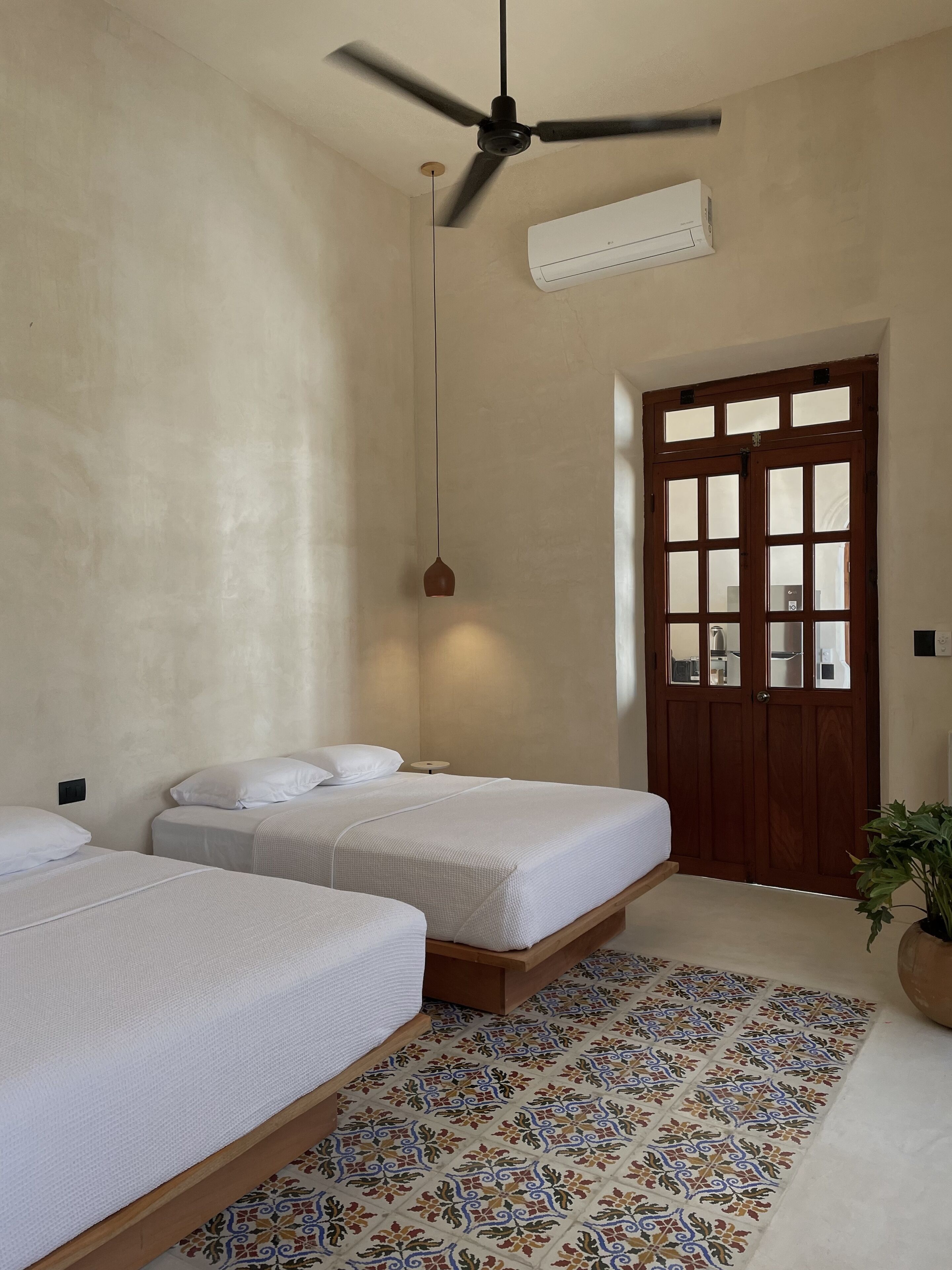 Una habitación con dos camas, ventilador de techo, aire acondicionado, puerta de madera con paneles de vidrio y piso con diseño.