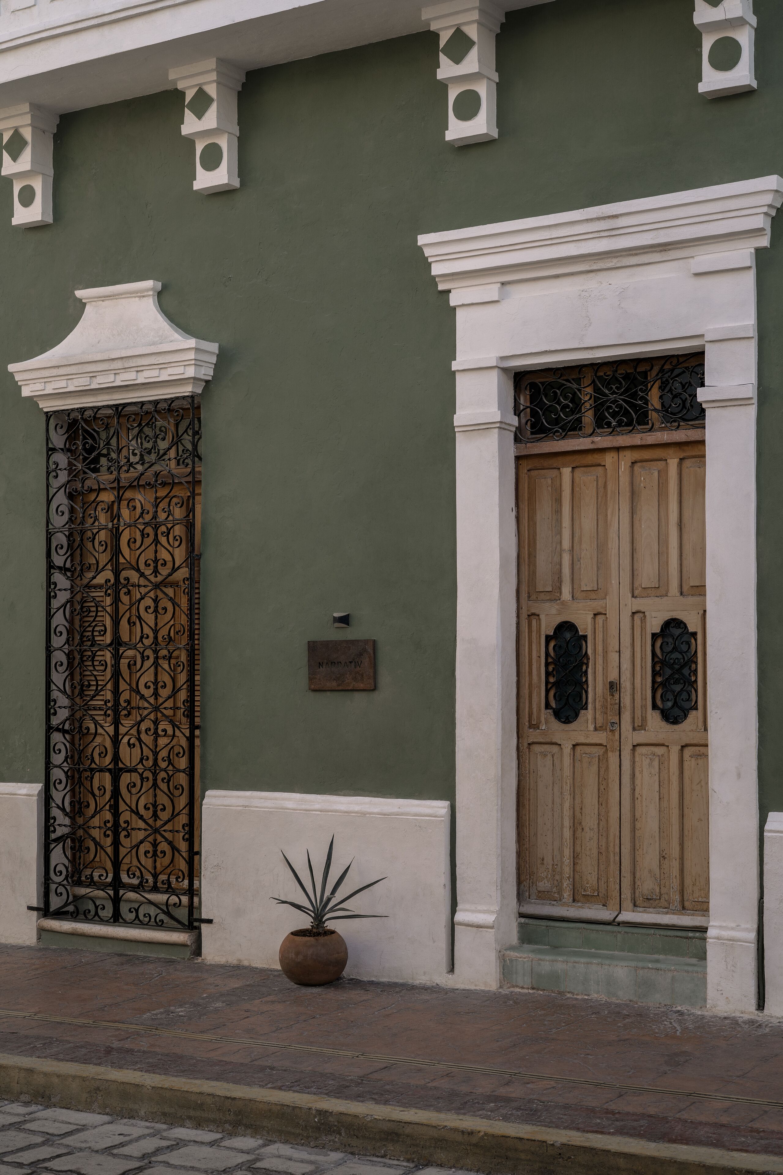 A building facade with a green wall, a wooden door with decorative ironwork, and a potted plant on the ground.