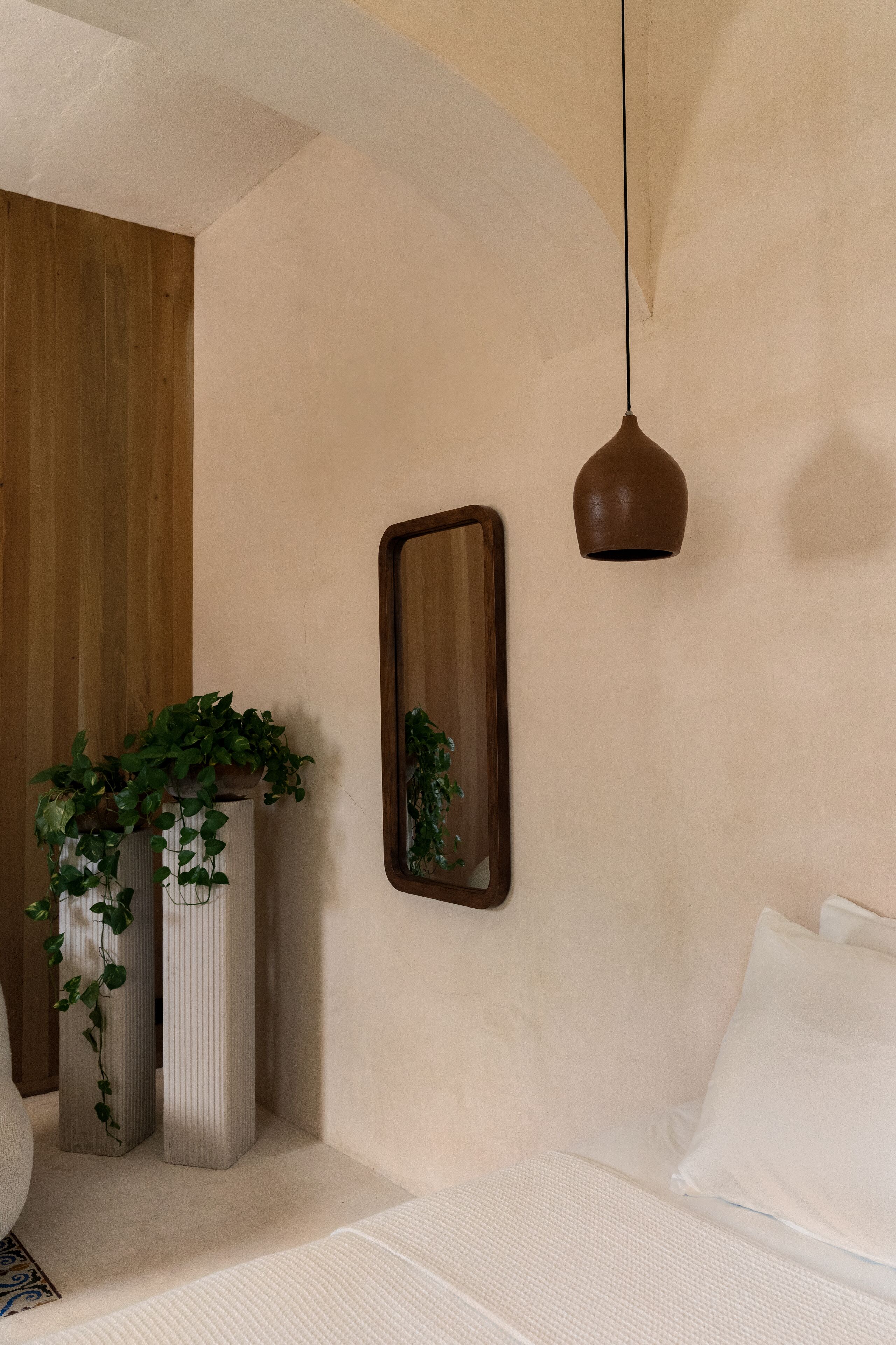 A bedroom with a bed, two tall vases with plants, a wooden panel wall, a mirror, and a hanging pendant light.