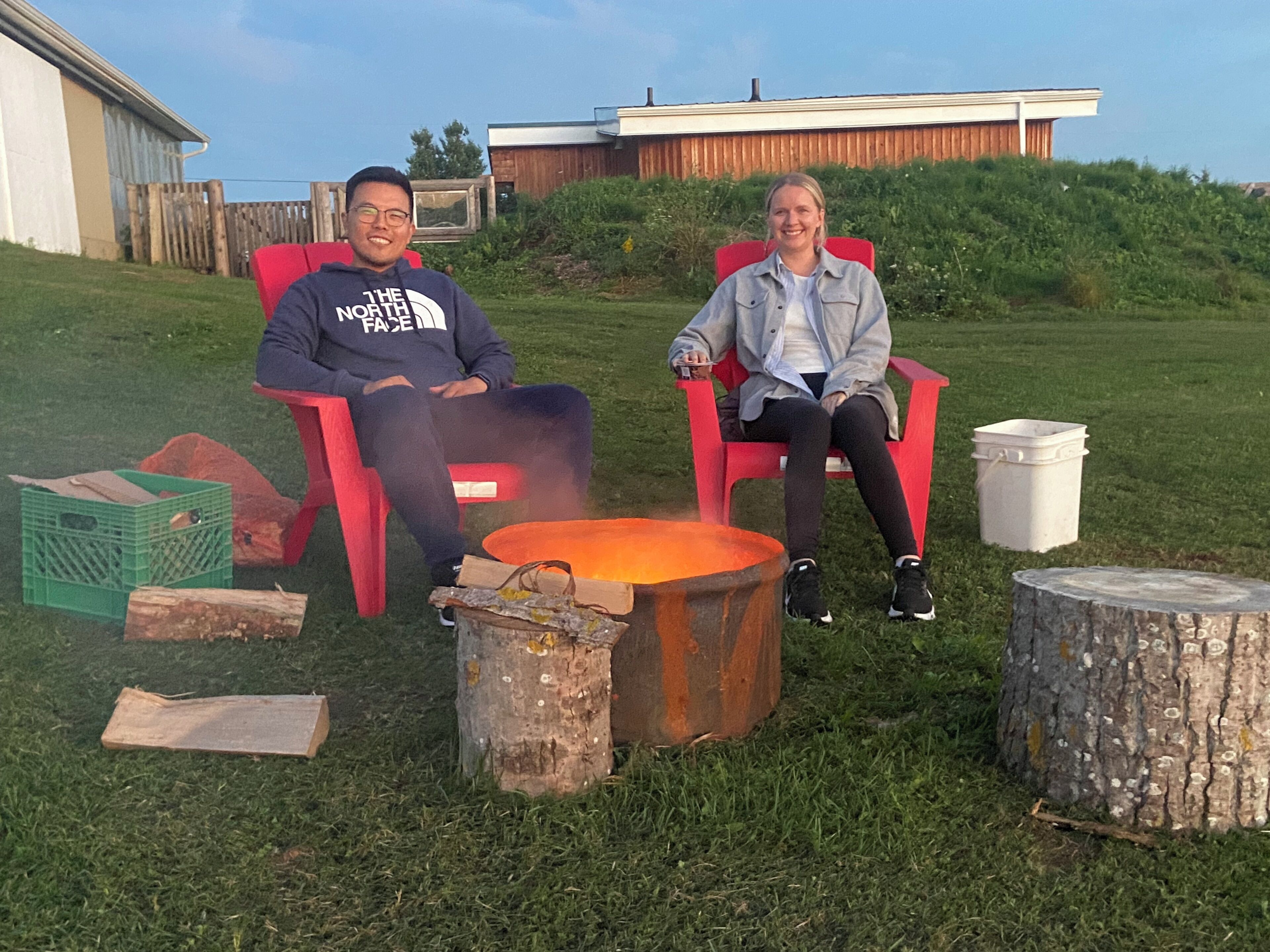Two people seated on red chairs outdoors with a fire pit in the foreground.