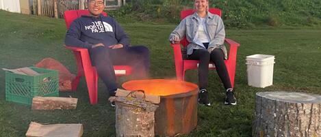 Two people seated on red chairs outdoors with a fire pit in the foreground.