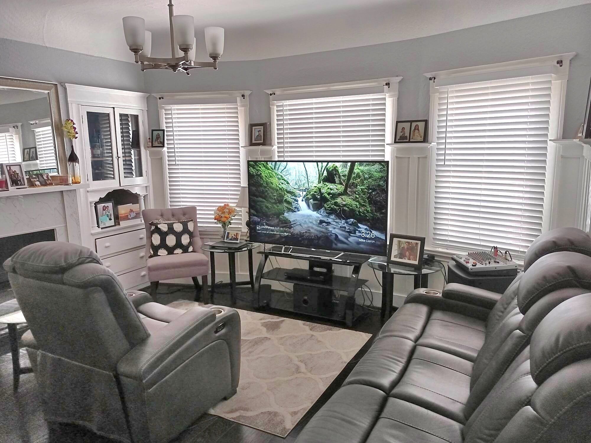 A living room with a large flat-screen TV, a sofa, a chair, and a coffee table. There are two windows with blinds, a chandelier, and a mirror on the wall.