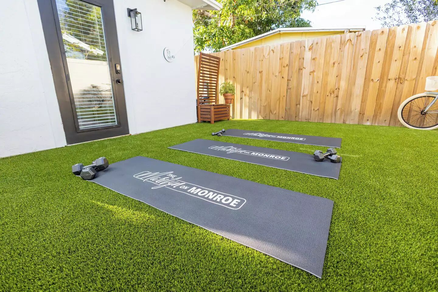 Deux tapis de sol pour exercices, posés sur une pelouse artificielle, avec des haltères, une clôture en bois et un vélo dans un jardin.