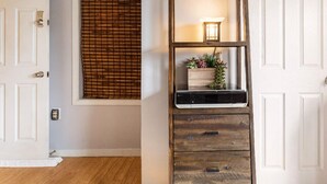 Une pièce avec un plancher en bois, une porte blanche, une fenêtre avec un store en bambou et une étagère en bois sur laquelle se trouvent une petite lampe et des plantes.