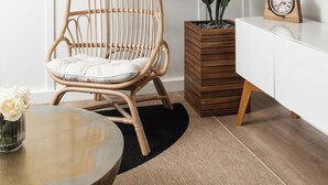 Una sala de estar moderna con una silla de ratán, una planta en maceta, una mesa redonda con un florero, una mesita de madera y un televisor montado en la pared.
