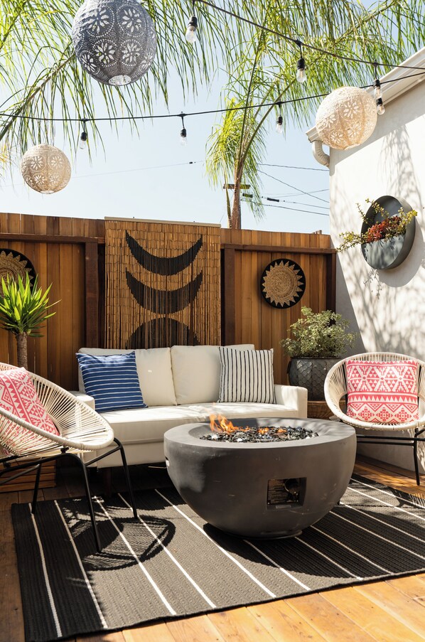 An outdoor patio with a sofa, chairs, a fire pit, and decorative elements like hanging lights and wall art.